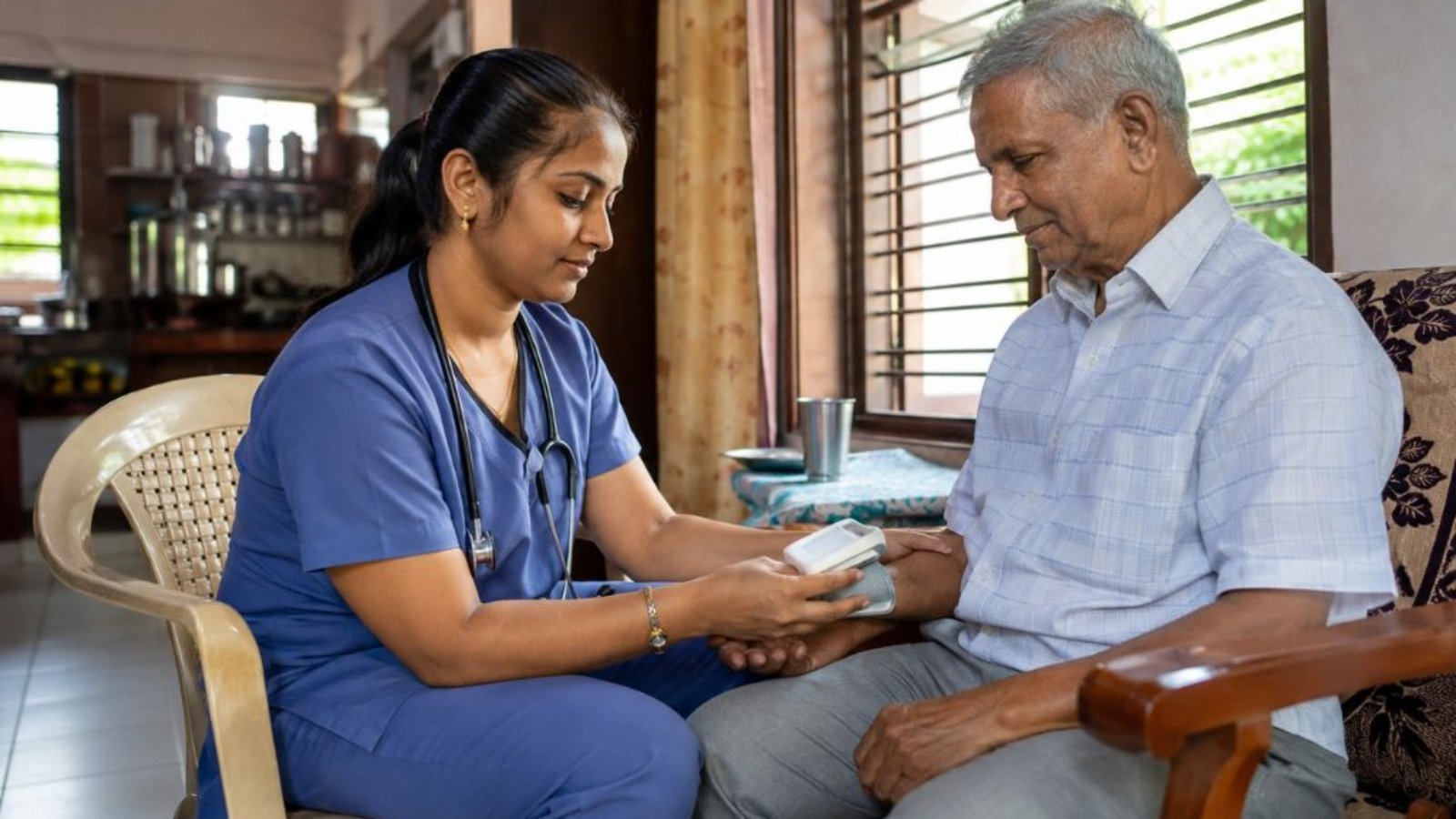 Home nurse in scrubs sitting beside an elderly man in his living room at home