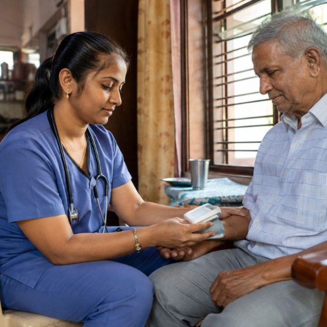 Home nurse in scrubs sitting beside an elderly man in his living room at home